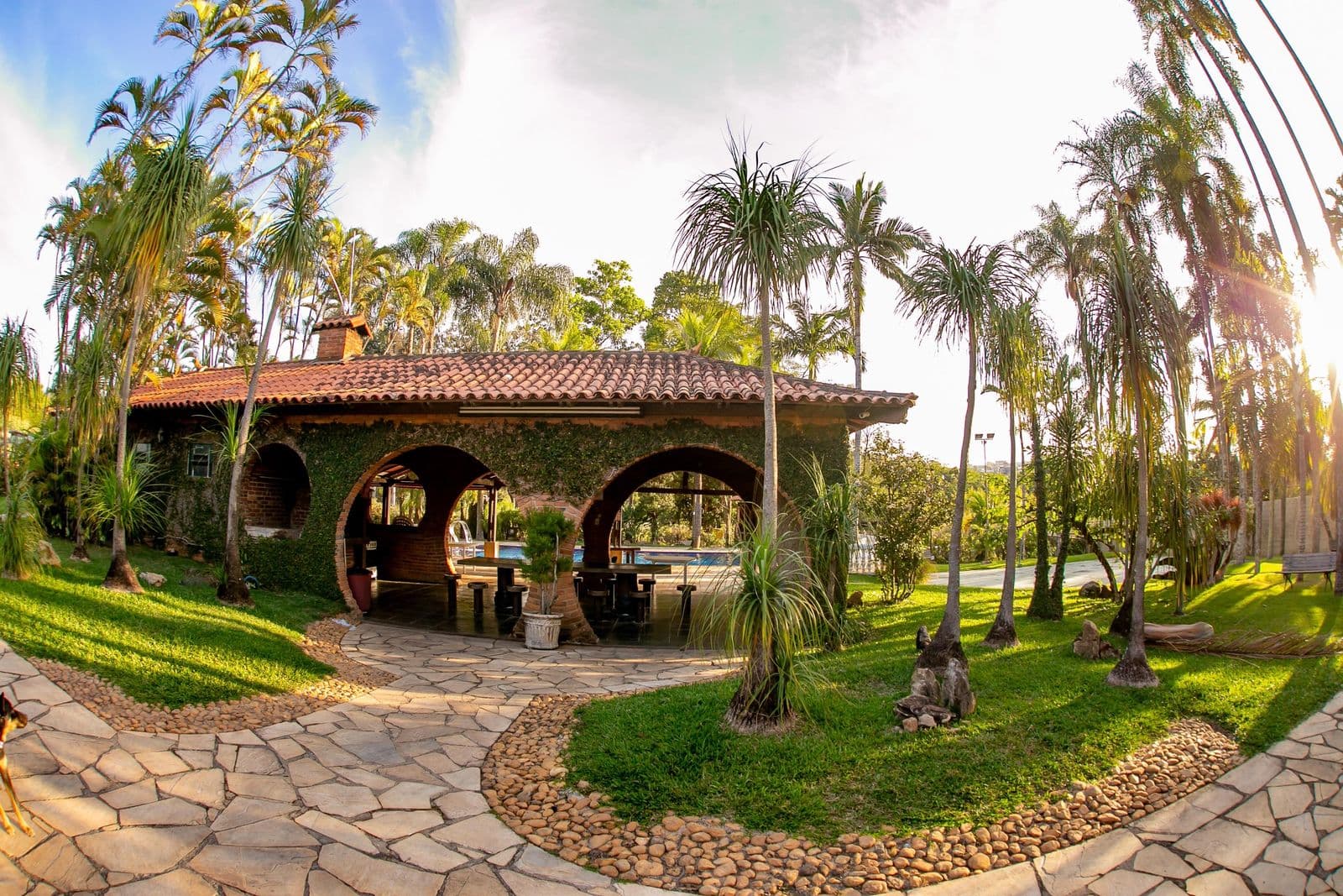 Quiosque de arcos de tijolo no fim da tarde, com o jardim e a piscina ao redor