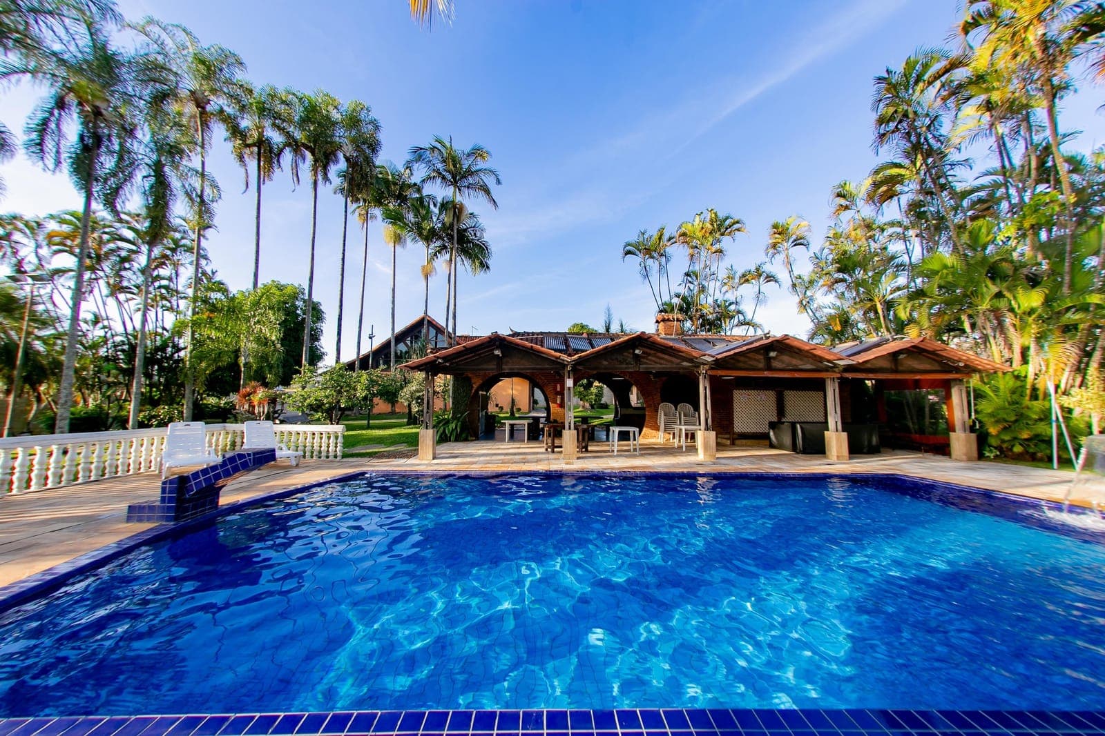 Piscina de azulejo azul cercada por palmeiras imperiais, com quiosques de tijolo ao fundo