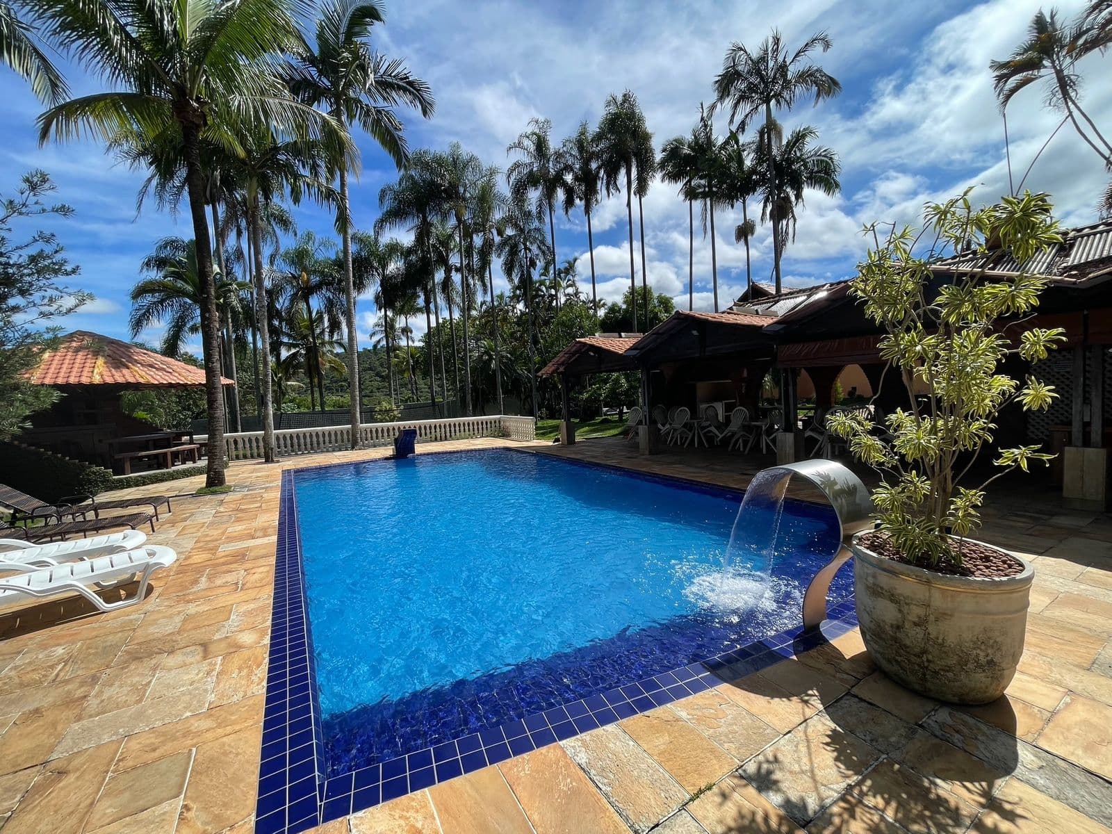 Piscina com a cascata ligada, vasos de planta no piso de pedra e os quiosques ao lado