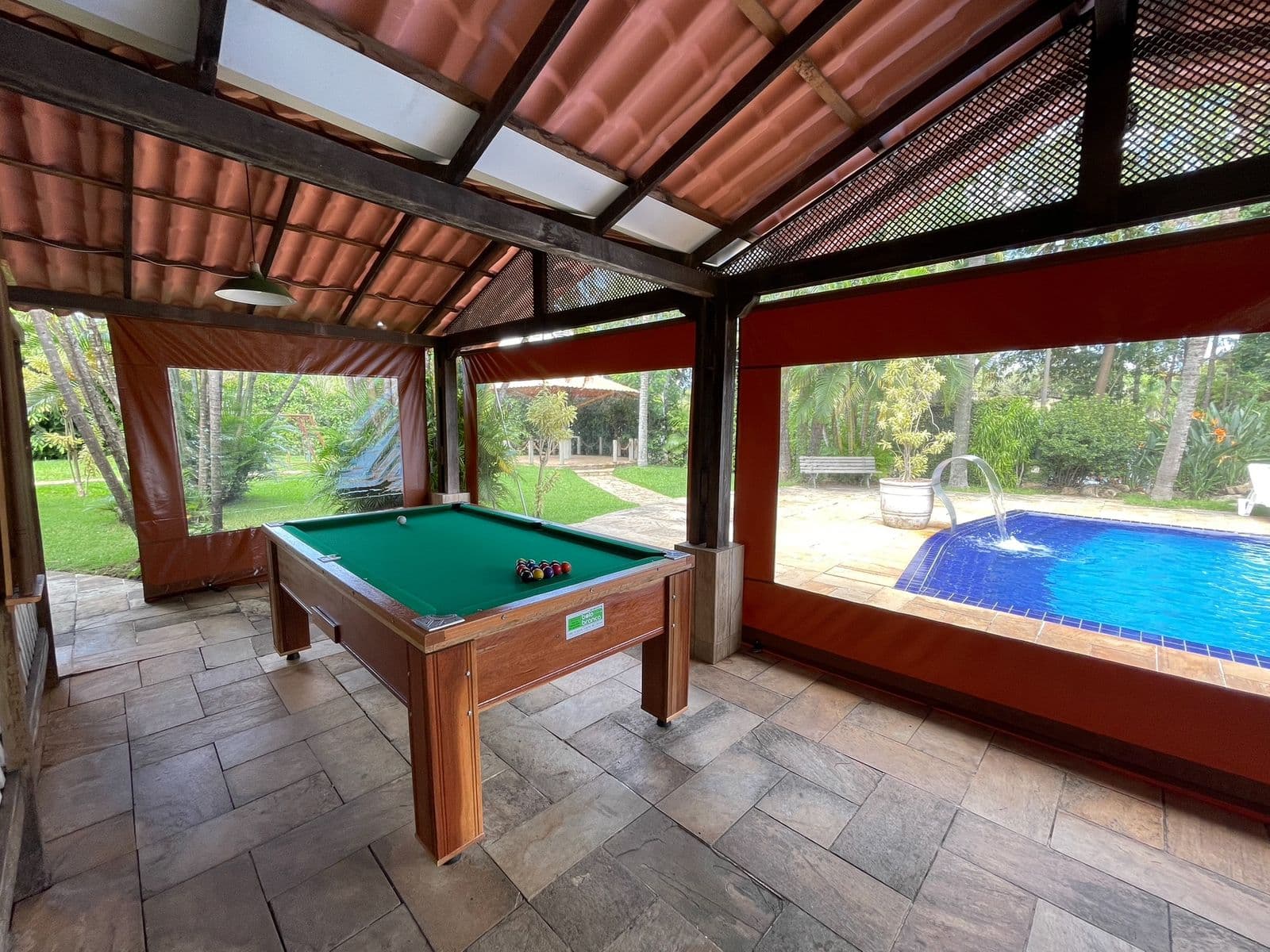 Mesa de sinuca sob o telhado de telha, com a piscina logo ao lado