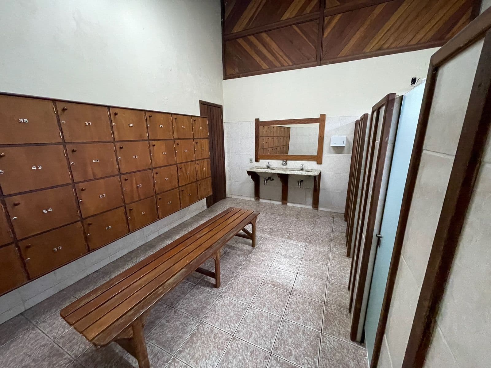 Vestiário com cabines, banco de madeira e pé-direito alto